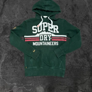 Grön Superdry hoodie med tryck - Mörkgrön hoodie från Superdry med stort vitt och rött tryck på bröstet där det står 'Super Dry Mountaineers'. Tröjan har huva med vita snören och känguruficka framtill. Klassisk streetwear-stil, perfekt för dig som gillar sportig look.