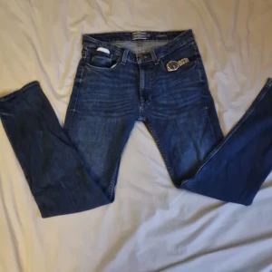 Blå jeans från Dressmann Regular Fit - Säljer ett par klassiska blå jeans från Dressmann i modellen Regular Fit. Jeansen har fem fickor, snygga kontrastsömmar och rak passform. Perfekta för dig som gillar en tidlös look och vill ha ett par bekväma jeans till vardags.