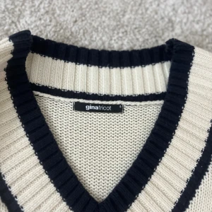 Vit stickad Tröja - Vit stickad tröja från Gina Tricot med marinblå ränder och bred v-ringning. Tröjan har ribbade muddar och detaljer vid hals, ärmslut och nederkant. Perfekt för en preppy look och passar till både jeans och kjol.