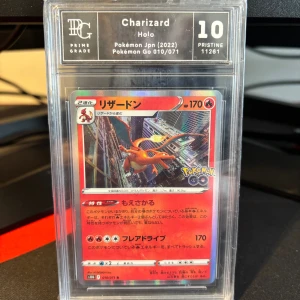 Charizard Holo Pokémon Go 010/071 - Upptäck den episka Charizard Holo-kortet från Pokémon Go-serien! Perfekt för samlare och Pokémon-fans som vill ha något unikt och coolt i sin samling. Kortet är i PRISTINE 10 skick och har en snygg design med Charizard i en stadsmiljö.