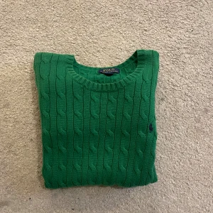 Grön kabelstickad tröja Polo Ralph Lauren - Snygg grön kabelstickad tröja från Polo Ralph Lauren med rund hals och diskret broderad logga på bröstet. Tröjan har långa ärmar och ribbade muddar vid hals, ärmslut och nederkant. Perfekt för dig som gillar klassisk stil med en modern touch.