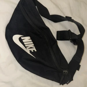 Svart axelväska från Nike - Svart axelväska från Nike med klassisk vit logga framtill. Väskan har justerbar rem och dragkedja upptill för smidig förvaring. Tillverkad i slitstark polyester, perfekt för att bära med sig det viktigaste. Enkel och stilren design som funkar till många outfits.