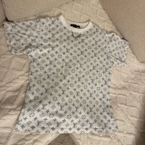 Vit Louis Vuitton t-shirt med kedjedetalj - Säljer en vit t-shirt från Louis Vuitton med grått monogramtryck över hela plagget. T-shirten har korta ärmar, rund halsringning och en cool silverfärgad kedja framtill vid halsen. Perfekt statement-piece för dig som gillar lyxiga streetwear-vibbar.