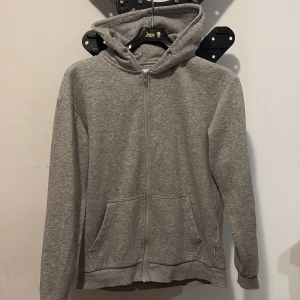 Grå hoodie från Jack & Jones - Grå hoodie från Jack & Jones med dragkedja framtill och två fickor. Mjuk insida i fleece och klassisk huva med snörhål. Perfekt för chill dagar och enkel att matcha med jeans eller joggers.