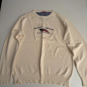 Beige stickad tröja från GANT - Snygg beige stickad tröja från GANT med broderad amerikansk flagga och texten 'GANT 1949 American Sportswear' på bröstet. Tröjan har rund hals och långa ärmar med ribbade muddar. Perfekt för dig som gillar klassisk och sportig stil.