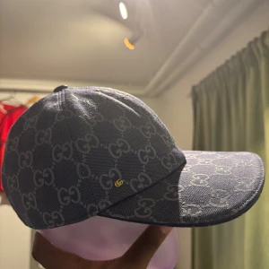Gucci - Snygg blå keps från Gucci med det klassiska GG-mönstret över hela kepsen. Kepsen har böjd skärm och en liten gul Gucci-logga på sidan. Materialet är bomull och polyester, insidan är ljus och har Gucci-etikett. Perfekt för dig som vill sticka ut med designerstil.