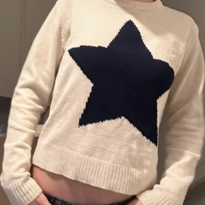 Stickad tröja  - Snygg stickad tröja med en stor marinblå stjärna framtill. Tröjan har rund hals, ribbade muddar och lång ärm. Perfekt för dig som gillar grafiska detaljer och vill ha en mysig, trendig look. Passar till jeans eller kjol för en cool stil.
