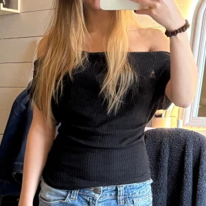 Svart ribbad offshoulder topp - Säljer en svart ribbad topp med offshoulder-design. Toppen är tight och har en snygg, croppad passform som framhäver axlarna. Perfekt att styla med jeans eller kjol för en cool och trendig look. Materialet är mjukt och följsamt.