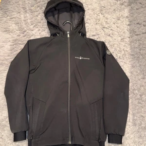 Sail Racing Softshell Hood jacket - Svart vindjacka från Sail Racing med huva och hel dragkedja framtill. Jackan har två sidofickor, diskret logga på bröstet och är tillverkad i ett slitstarkt syntetmaterial. Perfekt för blåsiga dagar och har en clean, sportig look. Storlek 160, passar xs. 😊