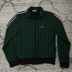 Grön mönstrad bomberjacka från Lacoste - Snygg grön bomberjacka från Lacoste med geometriskt mönster och klassisk krokodillogga på bröstet. Jackan har svarta muddar, vita ränder längs axlarna och hel dragkedja framtill. Insidan är mörkblå och materialet känns sportigt och bekvämt.