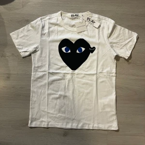 Vit Comme des Garçons Play t-shirt - Vit t-shirt med stort svart hjärta och blå ögon tryckt på bröstet. Klassisk rund hals och korta ärmar. Ikonisk design från Comme des Garçons Play, perfekt för en stilren och trendig look.