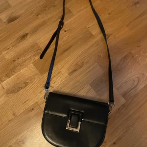 Svart axelväska med spänne - Stilren svart axelväska i skinnimitation med justerbar rem och snyggt metallspänne framtill. Remen går också att ta loss om man vill det. Väskan har flera fack inuti, inklusive ett med dragkedja, perfekt för att hålla ordning på dina grejer. Enkel och klassisk design som passar till många outfits. Bredd: 20cm höjd: 16cm.        Inte superkvalitet men funkar att använda ändå. Därför lågt pris