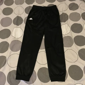 Svarta träningsbyxor från Kappa - Svarta träningsbyxor från Kappa med klassisk logga i vitt på vänster ben. Byxorna har elastisk midja och muddar vid bensluten för en skön passform. Materialet är glansigt syntet, perfekt för sport eller chill. Snygg och enkel design som funkar till streetwear.