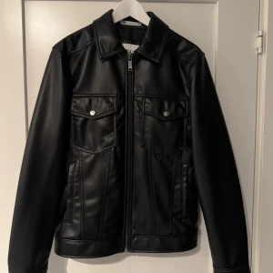 Svart skinnjacka från Zara - Snygg svart skinnjacka från Zara med klassisk krage, dragkedja framtill och två bröstfickor med knappar. Jackan har en stilren look och raka linjer, perfekt för dig som gillar minimalistisk stil. Materialet är slätt och ger en cool vibe.