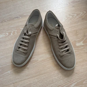 Beige mockasneakers Jim Rickey - Snygga beige sneakers från Jim Rickey i modellen Cloud, tillverkade i Portugal. Skorna har en stilren design med rund tå, platt vit sula och snörning. Utsidan är i mjuk mocka och insidan är bekvämt fodrad. Perfekta för dig som gillar minimalistisk stil.