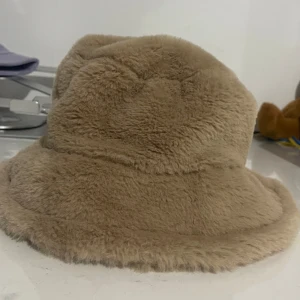 Beige fluffig bucket hat - Supermjuk och fluffig bucket hat i beige färg. Hatten har en klassisk form med bred brätte och är tillverkad i fuskpäls som ger en mysig och trendig look. Perfekt accessoar för att sticka ut och hålla värmen under kyliga dagar.