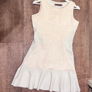 Vit kortklänning från Villa Hysé - Supersnygg vit kortklänning med swirl-mönster och volangkant nedtill. Klänningen är ärmlös och har en figurnära passform som framhäver siluetten. Materialet känns mjukt och har en lätt glans. Perfekt för dig som gillar stilrena detaljer och unik design. Snabb leverans 📦 