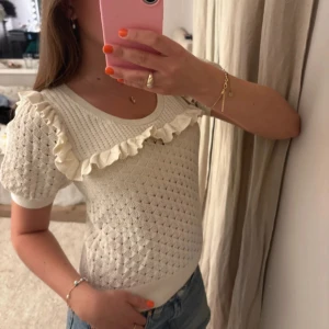 Vit stickad topp med volang - Supersöt vit stickad topp med korta ärmar och volangdetalj över axlarna. Mönstrad stickning ger en unik look och den mjuka ribbade kanten sitter snyggt i midjan. Perfekt att styla med jeans eller kjol för en fräsch vibe.