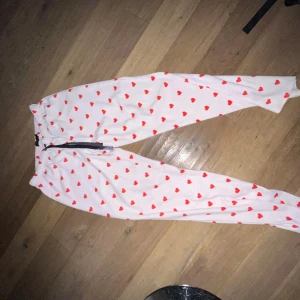 Vita mjukisbyxor med röda hjärtan - Supergulliga vita mjukisbyxor med röda hjärtan över hela tyget. Byxorna har resår i midjan och en svart dragsko för perfekt passform. Perfekta för mysiga dagar hemma eller när du vill ha något extra sött på dig.