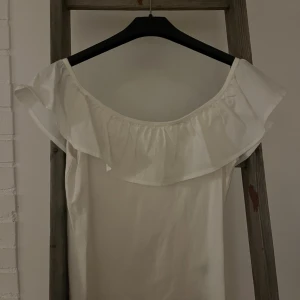 Vit offshoulder topp med volang - Säljer en söt vit offshoulder topp med bred volang runt halsen. Toppen är ärmlös och har en luftig passform, perfekt för varma dagar. Materialet känns mjukt och lätt, troligen bomull eller polyester. Enkel och stilren design som ger en romantisk vibe.