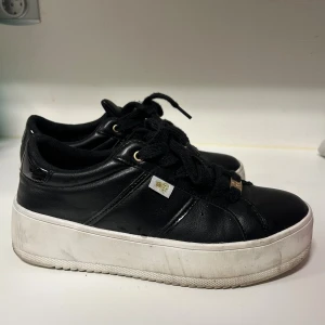 Svarta sneakers med platåsula från Pernilla Wahlgren  - Svarta sneakers med vit platåsula och snörning. Skorna har detaljer i guld på snörena och en liten vit etikett på sidan. Ovandelen är i skinn och bakpartiet har en blank finish. Snygg och chunky look som passar till streetwear. 40/41