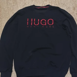 Svart sweatshirt från Hugo Boss - Svart sweatshirt från Hugo Boss med röd broderad logga på bröstet. Tröjan har rund halsringning, långa ärmar och ribbade muddar. Perfekt för dig som gillar stilren design med en tydlig logga framtill.