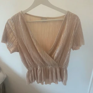 Champagnefärgad omlottopp med volang - Säljer en glansig champagnefärgad omlottopp med korta ärmar och v-ringning. Toppen har plisserat tyg, volangkant nedtill och ett snyggt omlottliv som ger en feminin siluett. Perfekt för dig som gillar att sticka ut med stilrena detaljer. brukade vara långärmad men klipptes till kort