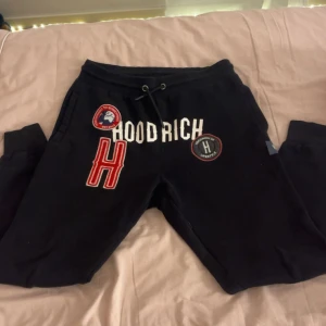 Svarta Hoodrich mjukisbyxor - Svarta mjukisbyxor från Hoodrich med coola patchar och broderade detaljer framtill. Byxorna har snörning i midjan, ribbade muddar vid bensluten och tryckt logga på baksidan. Perfekt för en avslappnad streetwear-look.