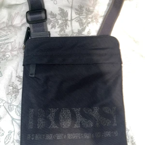 Svart axelväska från BOSS - Snygg svart axelväska från BOSS med justerbar rem och diskret logga framtill. Väskan har dragkedja upptill och ett innerfack med extra dragkedja. Tillverkad i slitstark polyester med stilren, rektangulär form. Perfekt för att bära med sig det viktigaste.