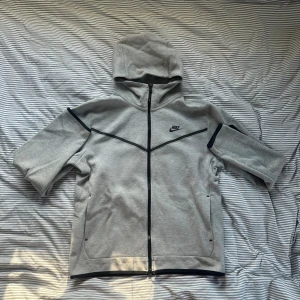 Grå Nike Texh strl L - Snygg grå tröja från Nike med svart logga på bröstet och svarta detaljer längs dragkedjan och ärmarna. Tröjan har huva, hel dragkedja och praktiska fickor, inklusive en med dragkedja på ärmen. Perfekt för dig som gillar sportig stil.