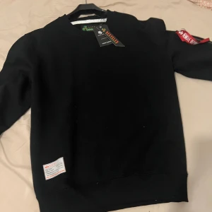 Helt ny Alpha Industries sweatshirt - Svart sweatshirt från Alpha Industries med klassisk passform. Tröjan har rund hals, lång ärm och en ikonisk röd tag på ärmen med dragkedja. Tillverkad i mjuk bomullsmix och har en liten patch nertill. Perfekt för en clean och stilren look. pris kan diskuteras.