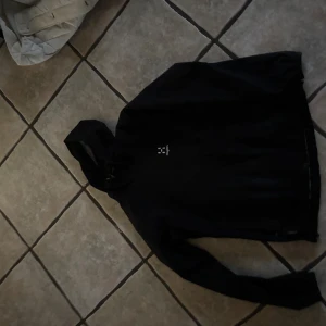 Svart vindjacka från Haglöfs - Svart vindjacka från Haglöfs med huva och dragkedja framtill. Jackan har en stilren design med logga på bröstet och långa ärmar. Perfekt för blåsiga dagar och outdoor-aktiviteter. Snygg och funktionell för dig som gillar friluftsliv.