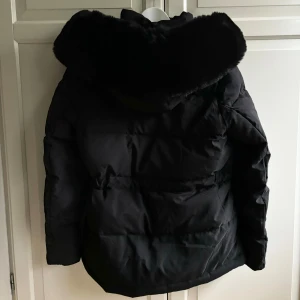 Svart vinterjacka med päls  - Knappt använd vinterjacka i väldigt fint skick. Pälsen på luvan är inte äkta. Justerbart midjebälte. Storlek L men passar även M