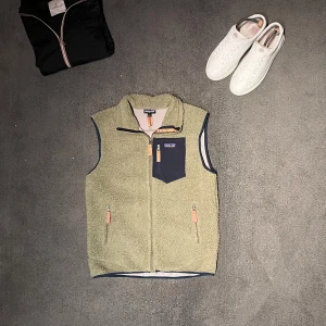 Patagonia fleece väst - Tjaaa säljer just nu en  fleeceväst från Patagonia med marinblå detaljer och dragkedjor. Västen har en hög krage, tre fickor med dragkedja och ett coolt bröstfack i marinblått. Västen är i mycket fint skick och är i storlek L men passar mig som har storlek M och är 182 och väger 69 kg. Jag är altid intresserad av byten om du bor i Stockholm.Har du några ytterligare frågor är det bara att höra av dig!!