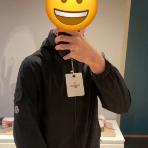 Svart vindjacka från Moncler - Svart vindjacka från Moncler med huva och dragkedja framtill. Jackan har Moncler-logga på ärmen och elastiska muddar vid handlederna. Materialet är lätt och syntetiskt, perfekt för blåsiga dagar. Snygg och stilren design med praktisk ficka på ärmen. Måtten:  Armar= 64cm Bredden=45cm  Längden= 65,5 cm 