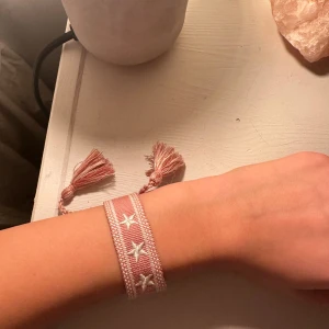 Rosa armband med stjärnor och tofsar från Spanien  - Ett brett, vävt armband i ljusrosa med vita broderade stjärnor och två tofsar i ändarna. Perfekt accessoar för att ge din outfit en söt och trendig touch.