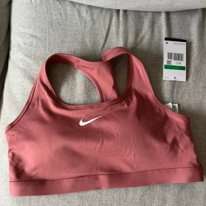 Rosa sporttopp från Nike i XL - Nike sporttopp i en snygg rosa färg med klassisk vit logga framtill. Modellen är ärmlös med brottarrygg och har en tight passform som sitter skönt vid träning. Materialet är mjukt och stretchigt polyester som andas bra. Perfekt för gymmet eller löprundan.