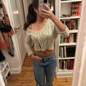 Beige croppad stickad kofta - Säljer en beige croppad stickad kofta/bolero med kabelstickat mönster och 2 knappar framtill. Koftan har lång ärm och en snygg v-ringad offshoulder-passform!