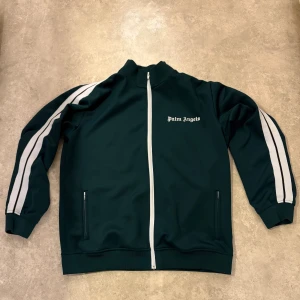 Palm Angels grön  - Palm Angels track jacket i mörkgrönt med vita ränder längs ärmarna och vit dragkedja framtill. Jackan har ståkrage, två fickor med dragkedja och logotyp på bröstet. Tillverkad i Italien i mjukt syntetmaterial. Perfekt för en sportig och clean look.