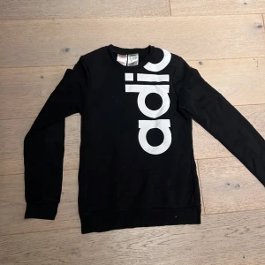 Svart Adidas tröja med stor logga - Svart långärmad tröja från Adidas med stor vit logga som går över axeln och ryggen. Tröjan har rund halsringning och är tillverkad i mjuk bomullsmix, perfekt för en sportig och avslappnad stil.