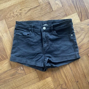 Svarta denimshorts från H&M - Svarta shorts från H&M i klassisk denimstil med fem fickor, uppvikta benslut och bälteshällor. Tillverkade i stretchig bomullsmix för en bekväm passform. Perfekta för varma dagar och enkel att matcha med allt.