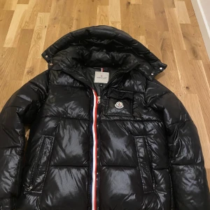 Svart M pufferjacka med huva - Scan funkar!!!Svart pufferjacka från M med glansig finish, avtagbar huva och ikonisk logga på bröstet. Jackan har en dragkedja med röd, vit och blå detalj, samt två sidofickor med dragkedja. Perfekt för kalla dagar och riktigt snygg stil.