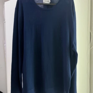 Mörkblå ulltröja från Calvin Klein - Snygg mörkblå rundhalsad tröja från Calvin Klein i mjuk och maskintvättbar ull. Enkel och stilren design med långa ärmar, perfekt för dig som gillar klassiska och bekväma plagg. Passar dig som vill ha en tidlös look med premiumkänsla.