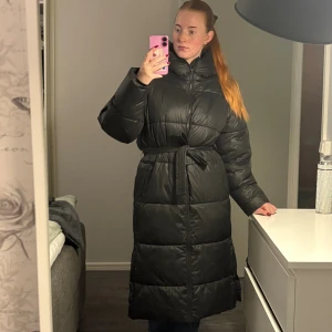 Svart lång pufferjacka med bälte - Säljer en svart, lång pufferjacka med huva och bälte i midjan. Super fin vinterjacka använd fåtal gånger 