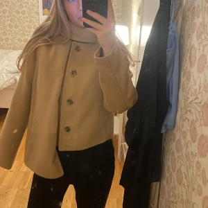 Beige kappa med inbyggd halsduk  - Säljer en beige kappa med en inbyggd halsduk, som också går att ta av och stora knappar framtill. Den är från HM. Pris kan diskuteras!!