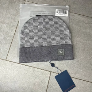 Grå rutig mössa  - Snygg grå mössa med klassiskt rutigt mönster. Mössan har uppvikt kant och är tillverkad i mjukt stickat material som känns lyxigt och bekvämt. Perfekt för dig som vill ha en stilren accessoar med designer-vibe.