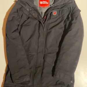 Svart parkajacka från Fjällräven XXS - Svart parkajacka från Fjällräven i storlek XXS. Jackan har huva, stora fickor med lock och dragkedja framtill. Klassisk Fjällräven-logga på bröstet och insidan. Perfekt för kalla dagar med sin långa modell och funktionella design.
