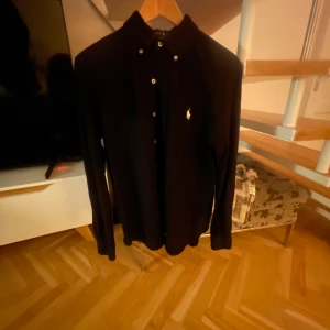 Mörkblå långärmad skjorta Ralph Lauren - Mörkblå långärmad skjorta från Ralph Lauren i modellen Featherweight Mesh. Klassisk krage med knappar framtill och den ikoniska logon broderad på bröstet. Skjortan har en stilren och tidlös look, perfekt för dig som gillar enkel och snygg design.