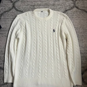 Vit stickad tröja från Ralph Lauren - Säljer en klassisk vit stickad tröja från Ralph Lauren med kabelstickat mönster och rund hals. Tröjan har långärmade ribbade muddar och den ikoniska Polo-loggan broderad på bröstet. Perfekt för dig som gillar stilren och tidlös design.
