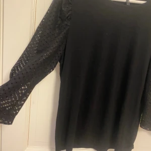 Svart blus med spetsärmar - Svart blus med rund hals och långa, transparenta spetsärmar med hålmönster. Ärmarna har en lätt puffig form och ger en cool kontrast mot den släta kroppen. Perfekt för dig som gillar stilrena plagg med en twist.